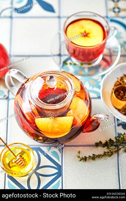 Fruit tea with apples and thyme in glass teapot and cup on table made of colored tiles