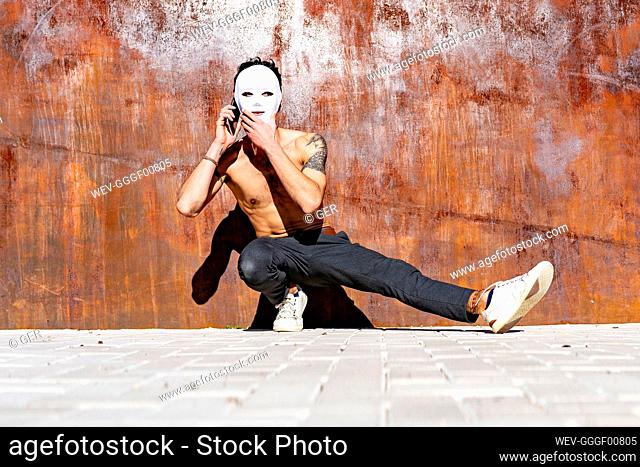 Young shirtless man wearing white mask talking on mobile phone while crouching against brown wall