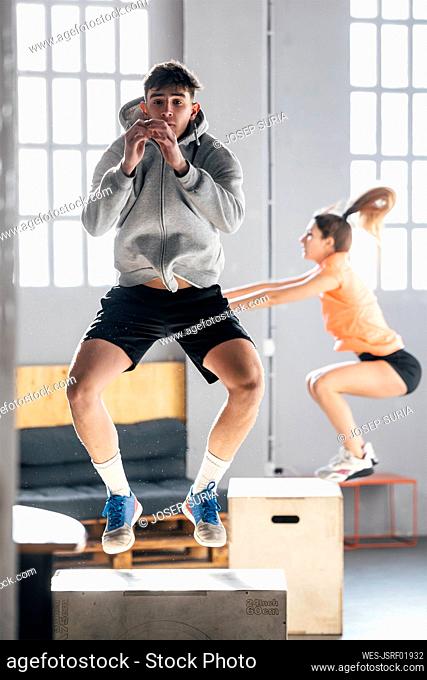 Young man with woman doing jumping squats on wooden box in gym
