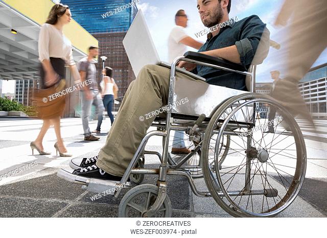 Man in a wheelchair working on his laptop between a crowd of people in a city