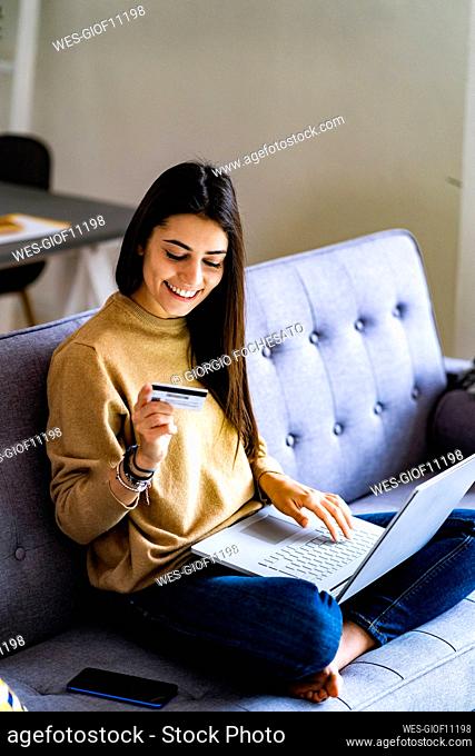 Smiling woman with credit card shopping online while sitting on sofa at home