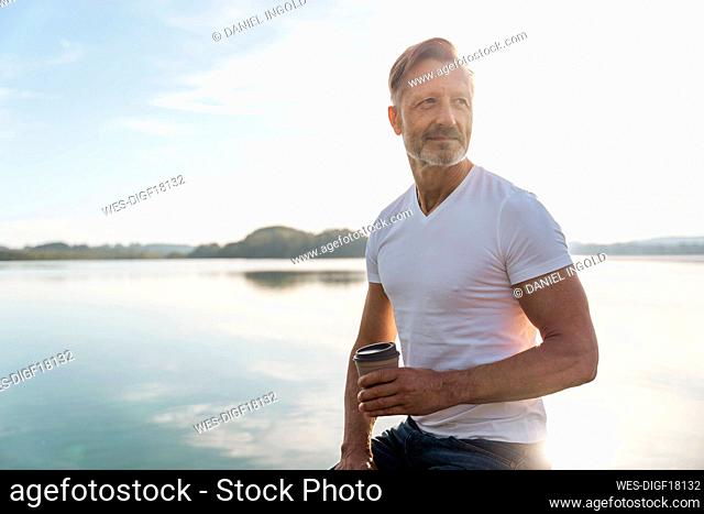Thoughtful mature man sitting with coffee cup near lake on sunny day