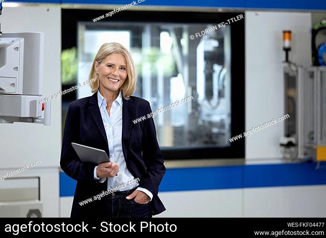 Mature businesswoman with hand in pocket holding digital tablet in factory