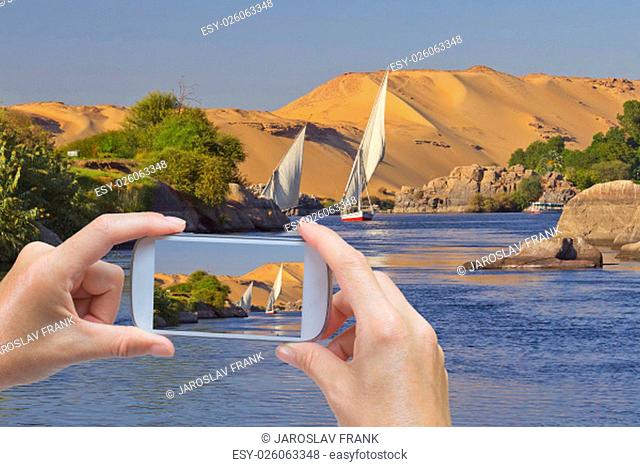In the bottom left of the photo are hands holding smart phone and taking picture of a sailing boat sailing the river Nile near egyptian city Aswan