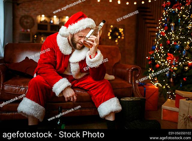 Bad Santa claus with bottle of alcohol sitting on couch, hangover. Unhealthy lifestyle, bearded man in holiday costume, new year and alcoholism
