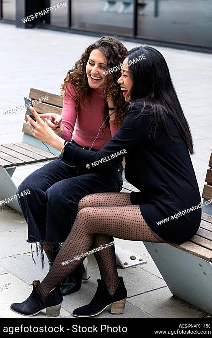 Women laughing while using mobile phone sitting on bench