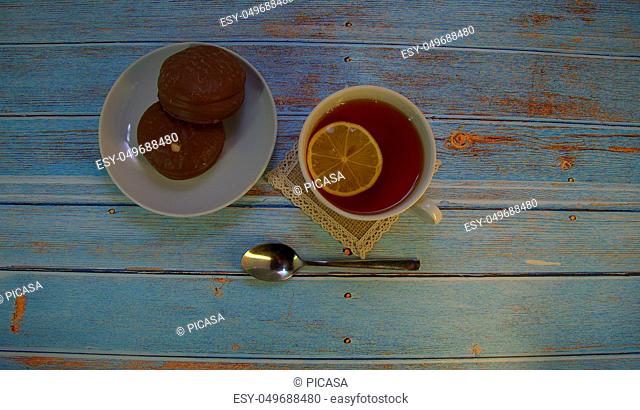 A cup of tea with lemon, a spoon and two chocolate cakes on a plate lie on a wooden table. Close-up