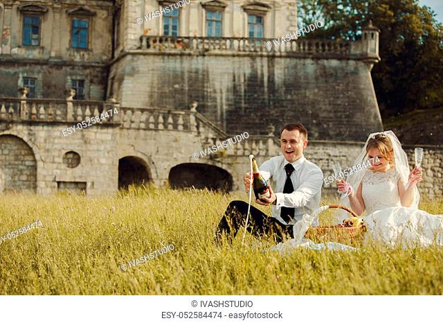 Groom opens a bottle of champagne sitting with a bride in the front of an old castle