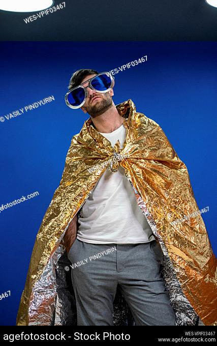 Male entrepreneur in novelty glasses wearing gold color wrapping paper cape against blue wall at work place