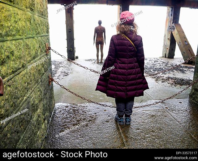 View taken from behind of a woman posing as Antony Gormley's cast iron figure, Another Time 1999-2013, looking out at the sea from the Folkestone Harbour Arm;...