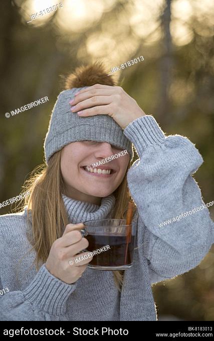 Portrait of a young woman in winter with cap, laughing, warming herself with a cup of tea, in the forest, Upper Bavaria, Bavaria, Germany, Europe