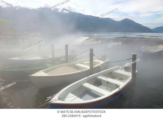 Small port on Alpine lake Maggiore with boats in the fog and Swiss Alps in Ascona, Switzerland