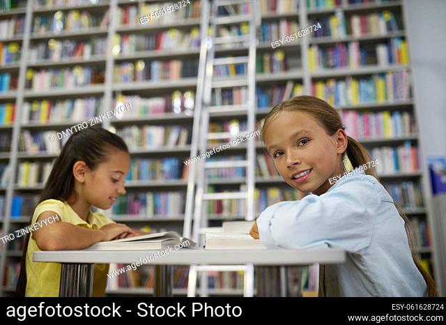Smiling pleased schoolgirl and her focused friend sitting at the table in the school library