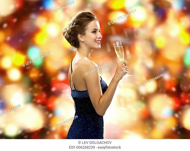 drinks, winter holidays, luxury and celebration concept - smiling woman in evening dress with glass of sparkling wine over red christmas lights background