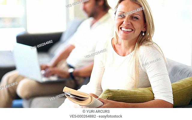 Mature woman reading on the sofa at home while her husband sits working in the background
