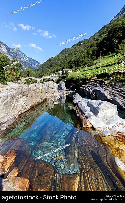 Switzerland, Ticino, Lavertezzo, Verzasca river flowing through Valle Verzasca in summer with Ponte dei Salti arch bridge in background