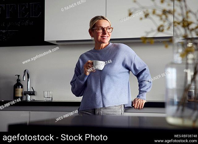 Smiling woman holding coffee cup leaning on kitchen counter