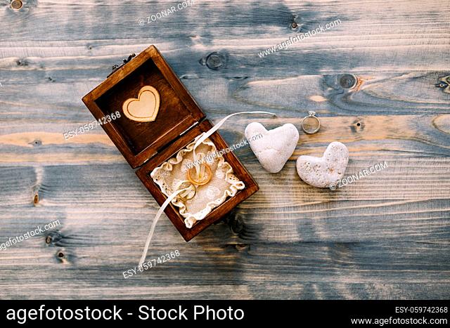 An open wooden box with wedding rings and two heart-shaped pebbles on a gray wood background. High quality photo