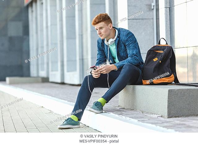 Redheaded young man sitting outdoors with smartphone and headphones