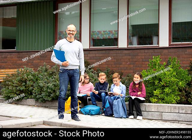 Smiling teacher in front of school, schoolchildren in background