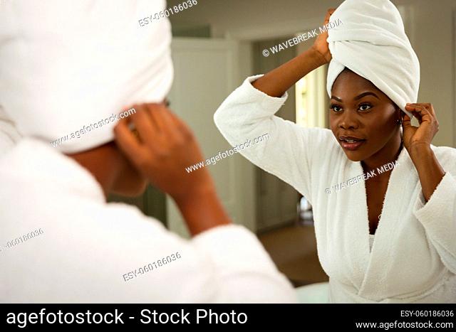 African american woman in bathroom wearing bathrobe, looking in mirror putting towel on head