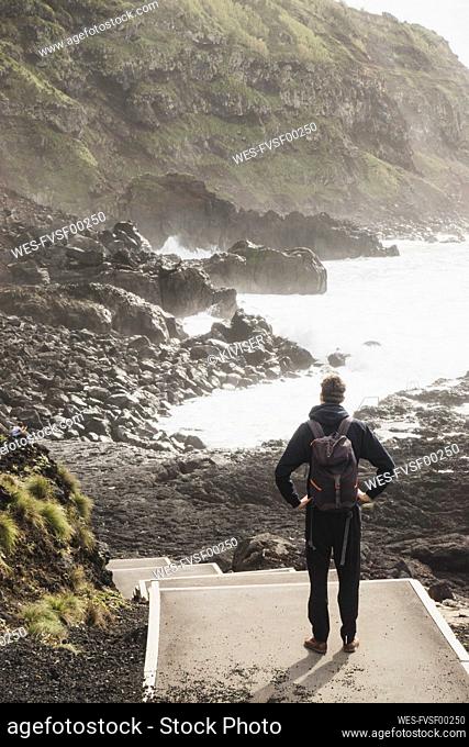 Full length rear view of man looking at rocky coastline at Ponta da Ferraria, San Miguel, Azores, Portugal