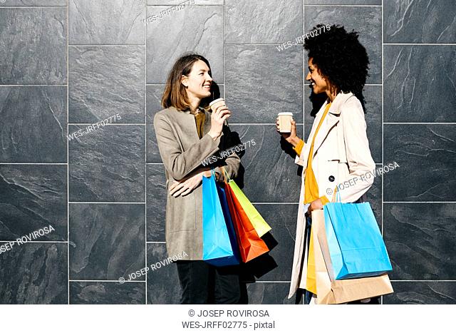 Two happy women with shopping bags and takeaway coffee standing at a wall talking