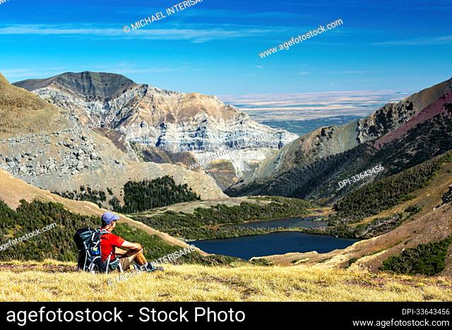 Male hiker relaxing on grassy mountain ridge overlooking an alpine lake, mountain ranges, blue sky and clouds in the background
