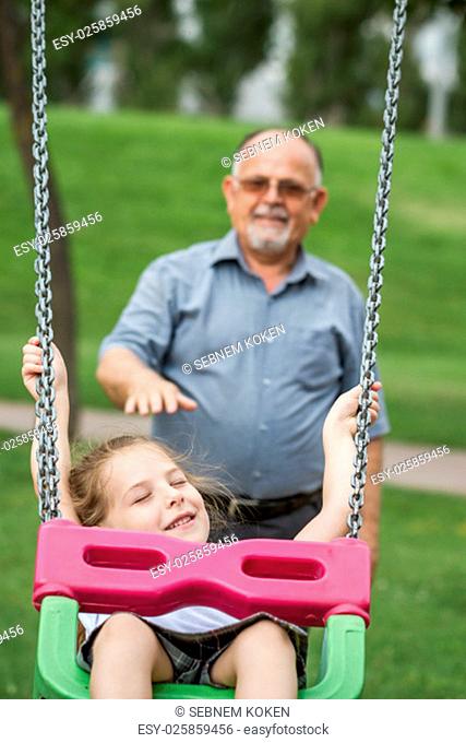 Little girl with her grandfather having fun on a swing in a green park