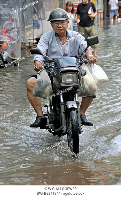 old man riding on a moped with full shopping bags through a street flooded after a heavy shower, Thailand, Bangkok