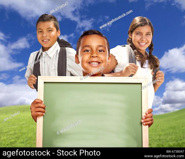 Smiling happy hispanic boys and girl in grass field holding blank chalk board