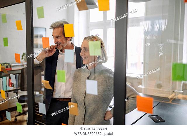 Happy colleagues looking at sticky notes at glass pane in office