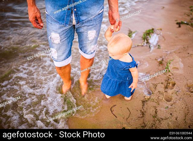 Dad teaches walks his little daughter holding his hand on a sandy beach near the sea. Father and daughter walk barefoot along the coastline