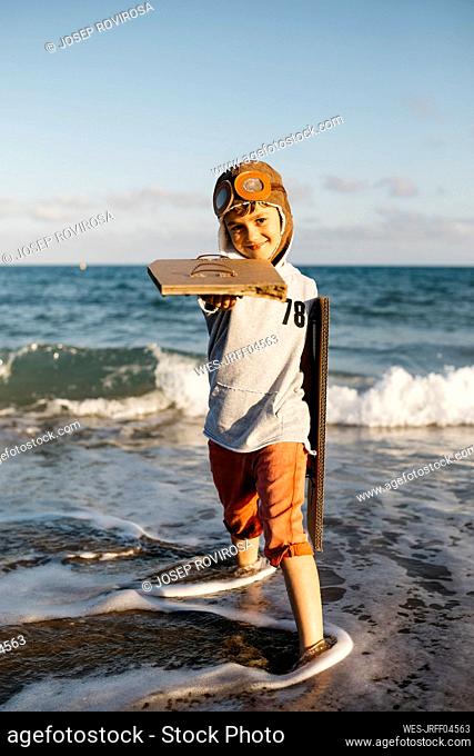 Boy with aviator's cap holding cardboard wings while standing at beach