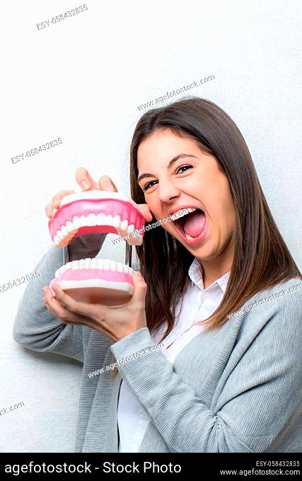Close up fun portrait of attractive young girl holding oversize human teeth prosthesis.Woman with open mouth against light textured background