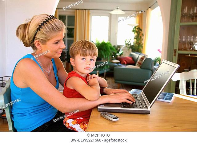 young mother with son, sitting at a table, using a laptop, a mobile lies on the table. The boy looks bored