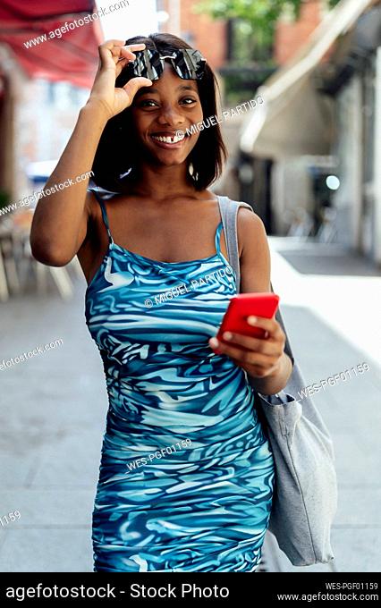 Happy woman wearing sunglasses with smart phone walking on road