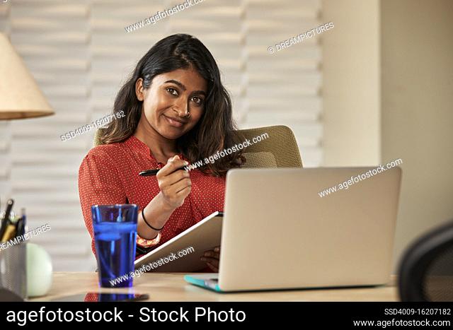 portrait of young ethnic woman sitting at desk with laptop computer taking notes on notepad