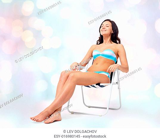 people, tanning, summer and beach concept - happy young woman in bikini swimsuit sunbathing on folding chair over blue holidays lights background