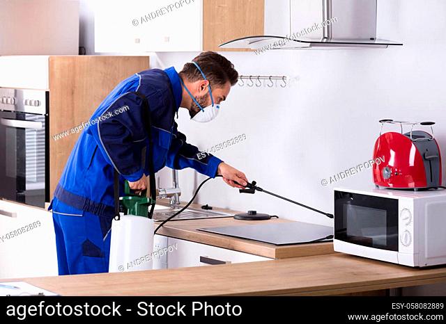 Pest Control Worker In Uniform Spraying Pesticide With Sprayer In Kitchen