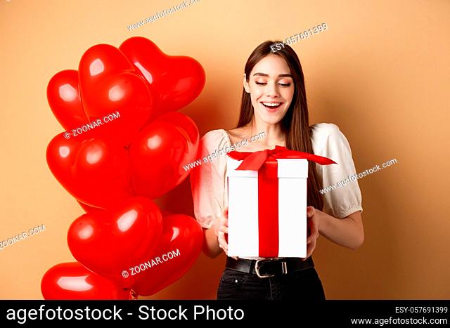 Happy woman open Valentines day gift, smiling excited and looking at present box, standing near red heart balloons on beige background