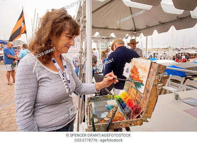 A Caucasian woman marine artist works at her easel in a Newport Beach, CA, yacht club boat yard depicting a vessel in the harbor. Note colorful paints