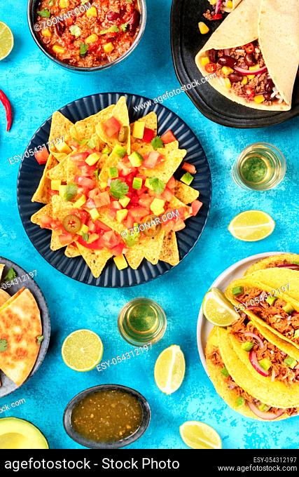 Mexican food, many dishes of the cuisine of Mexico, flat lay, top shot on a blue background. Nachos, tequila, taco shells, burritos, chili con carne