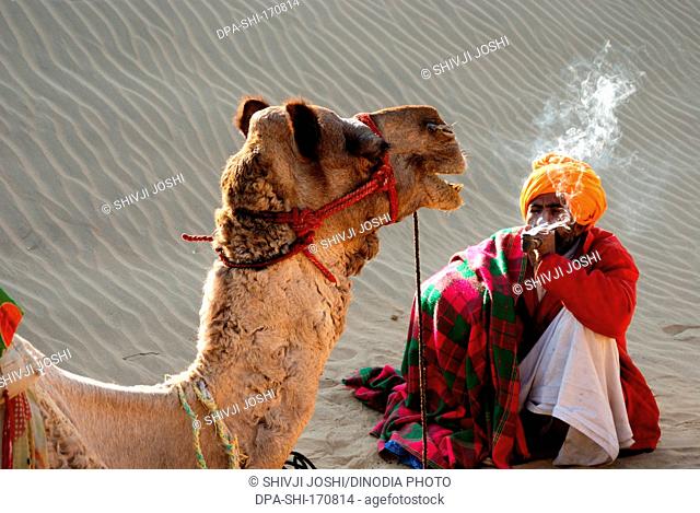 Camel and man smoking in desert in Khuri Khuhri ; Jaisalmer ; Rajasthan ; India NOMR