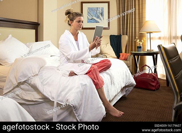 Young Caucasian woman wearing bathrobe in a hotel room, sitting on edge of bed using tablet device to connect with doctor for a video consultation