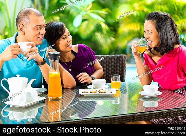 Asian friends having coffee on the porch in front of a home, in the background a tropical garden