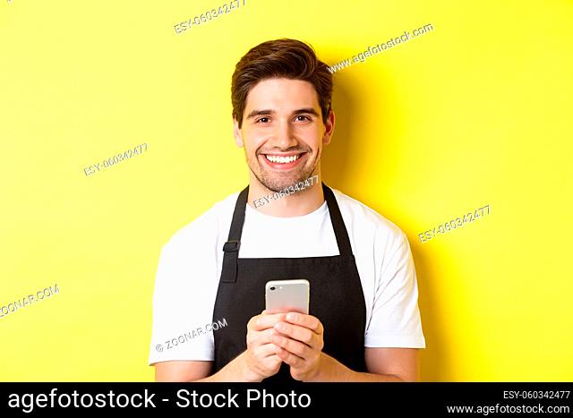 Close-up of handsome barista sending message on mobile phone, smiling happy, standing over yellow background