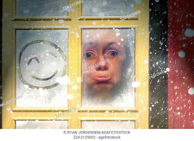 Sad woman stuck indoors by a window during a winter snow storm with happy expression squiggled on the glass. Trapped in boredom