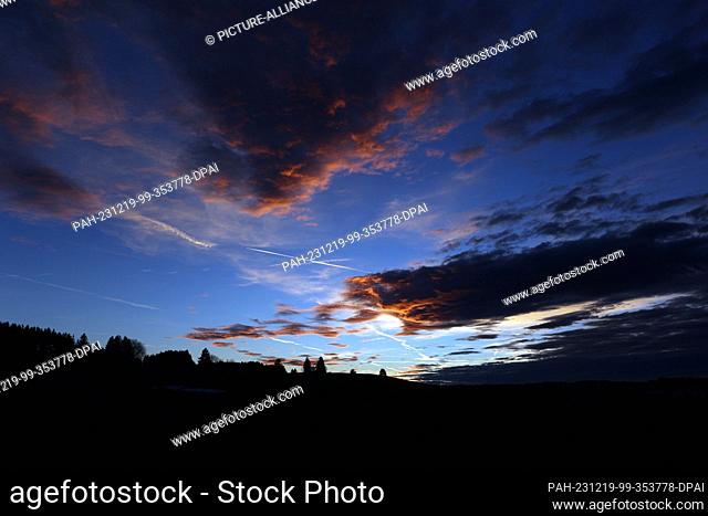 19 December 2023, Bavaria, Aitrang: The setting sun colors the clouds over the Allgäu landscape in different shades of red