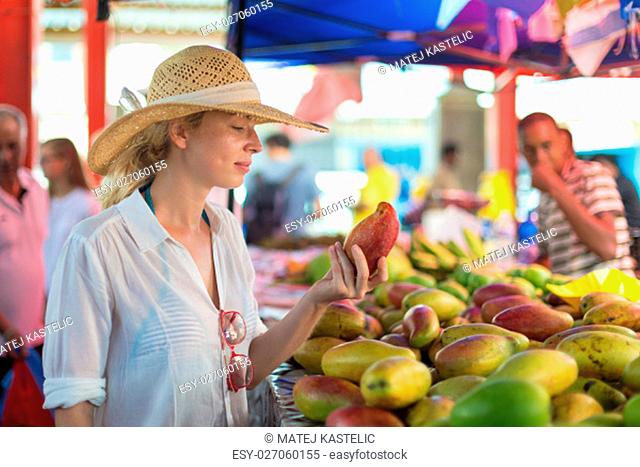 Female traveler wearing elegant colonial style white tunic and hat buying fresh tropical fruit on traditional Victoria food market on Seychelles islands